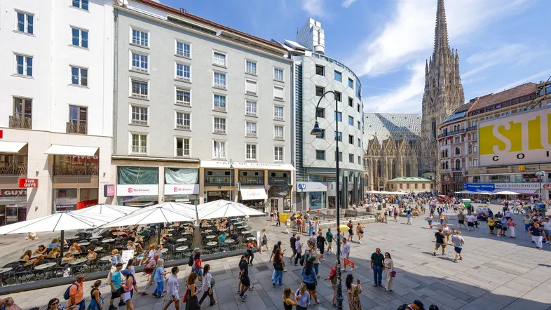 Belebter Stadtplatz mit historischen Gebäuden, moderner Architektur und dem Stephansdom im Hintergrund an einem sonnigen Tag.