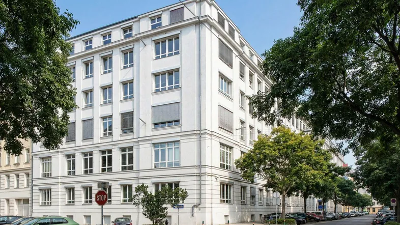 Weißes Bürogebäude mit vielen Fenstern und Jalousien an einer von Bäumen gesäumten Straße.