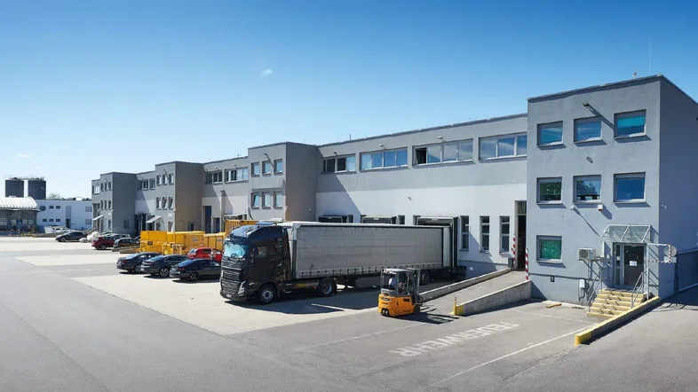 Großes Gewerbegebäude mit mehreren Laderampen, LKW-Parkplätzen und einem geräumigen Vorplatz unter blauem Himmel.