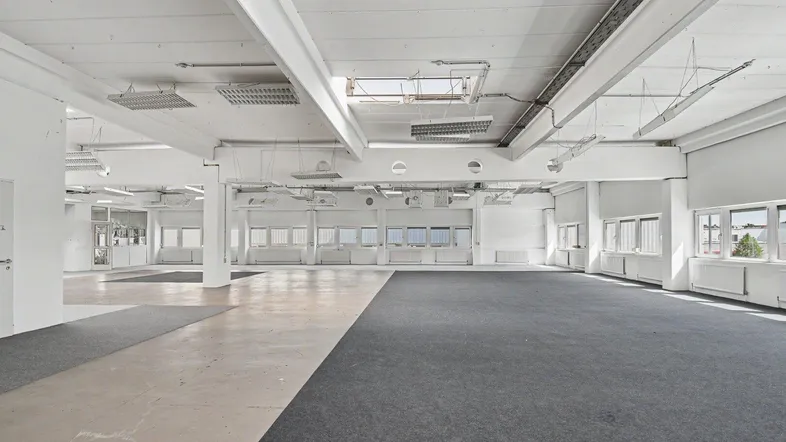 Großer, leerer Büroraum mit weißen Wänden, sichtbaren Deckenbalken und einer Kombination aus Teppich- und Betonboden.