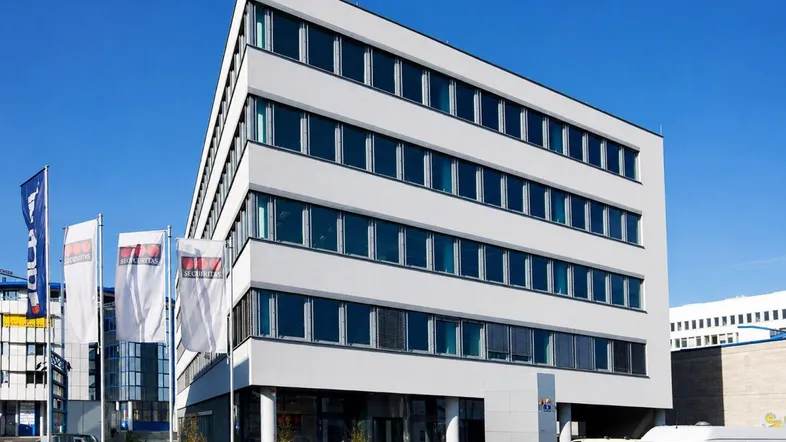 Modernes Bürogebäude mit heller Fassade, großen Fenstern und Firmenfahnen unter klarem blauem Himmel.