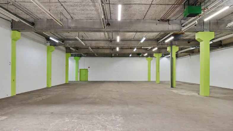 Großer, leerer Industrieraum mit Betonboden, sichtbaren Leitungen und leuchtend grünen Säulen.