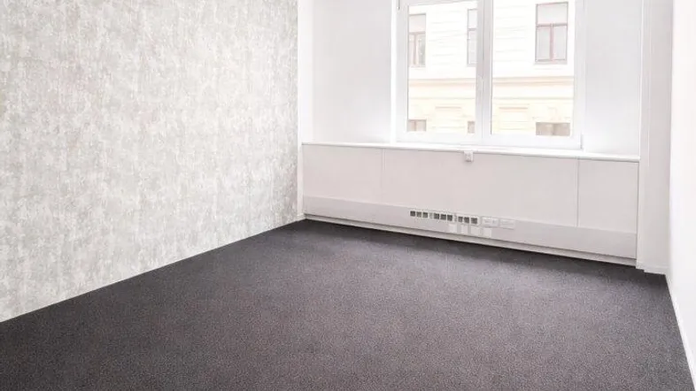 Leerer Büroraum mit dunklem Teppichboden, einer strukturierten Wand und großen Fenstern.