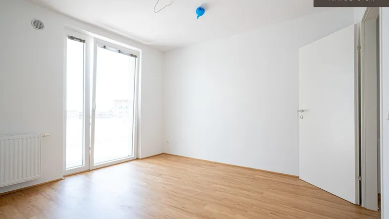Helles, leeres Zimmer mit Holzboden, weißen Wänden und Zugang zu einem Balkon oder einer Terrasse.