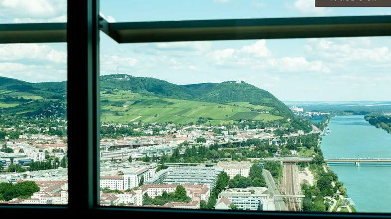 Blick aus einem Fenster auf eine weite Landschaft mit Stadt, Fluss und grünen Hügeln unter blauem Himmel.