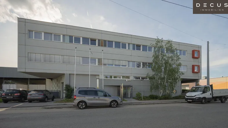 Modernes Bürogebäude mit grauer Fassade und roten Akzenten, umgeben von Parkplätzen und Bäumen.