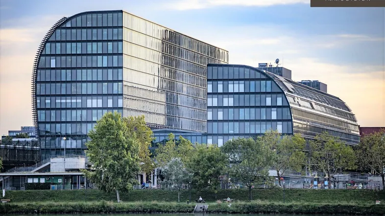 Modernes Bürogebäude mit geschwungener Glasfassade und Blick auf den Fluss, umgeben von Bäumen.