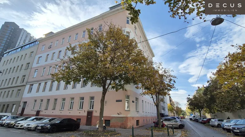Gebäudeansicht eines Mehrfamilienhauses an einer Straßenecke mit herbstlicher Bepflanzung und parkenden Autos.