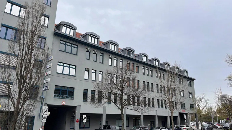 Graues Bürogebäude mit vielen Fenstern und geparkten Autos entlang der Straße unter bewölktem Himmel.