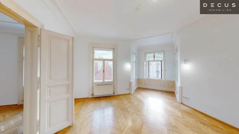Helles Wohnzimmer mit Fischgrätparkett, hohen Decken und Stuckverzierungen, durchflutet von Tageslicht.