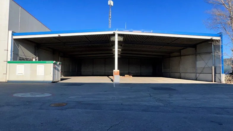 Große, offene Industriehalle mit blauer Dachkonstruktion und asphaltiertem Vorplatz unter klarem Himmel.