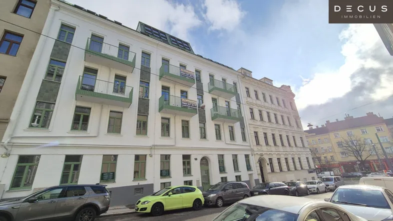 Klassisches Mehrfamilienhaus mit weißer Fassade, grünen Balkonen und parkenden Autos auf der Straße.