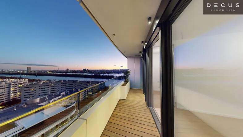 Geräumiger Balkon mit Holzboden, Glasgeländer und weitem Blick über die Stadt und den Fluss bei Sonnenuntergang.