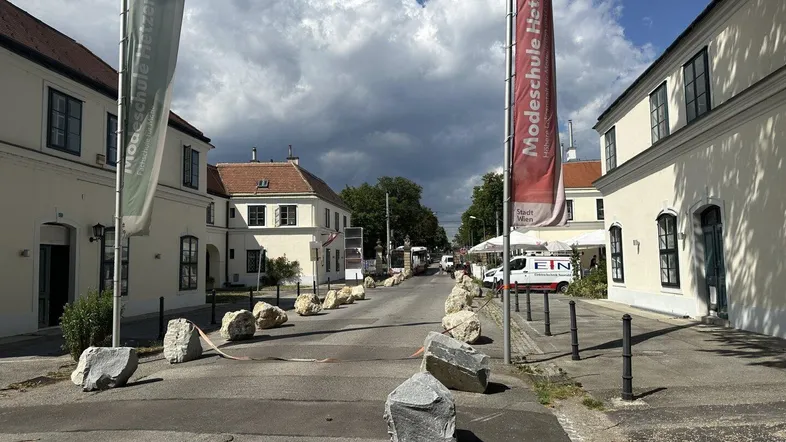 Eine Straße zwischen historischen Gebäuden mit großen Bannern und Felsbrocken als Absperrung unter bewölktem Himmel.