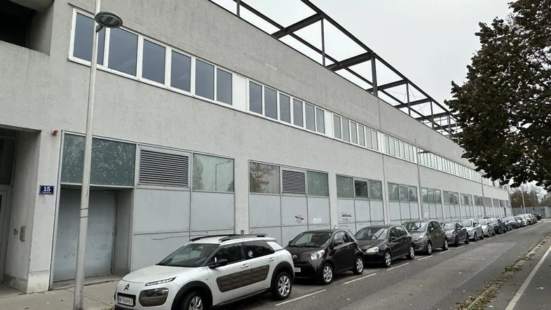 Lange Fassade eines Industriegebäudes mit parkenden Autos entlang der Straße unter bewölktem Himmel.