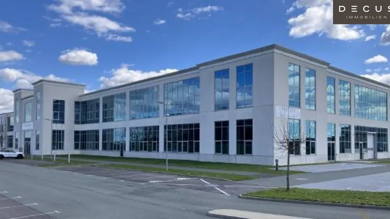 Modernes Bürogebäude mit großer Glasfassade und Parkplätzen unter blauem Himmel.