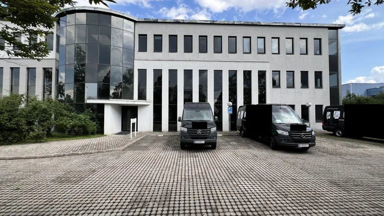 Modernes Bürogebäude mit Glasfassade und großem Parkplatz für Fahrzeuge, umgeben von Bäumen.
