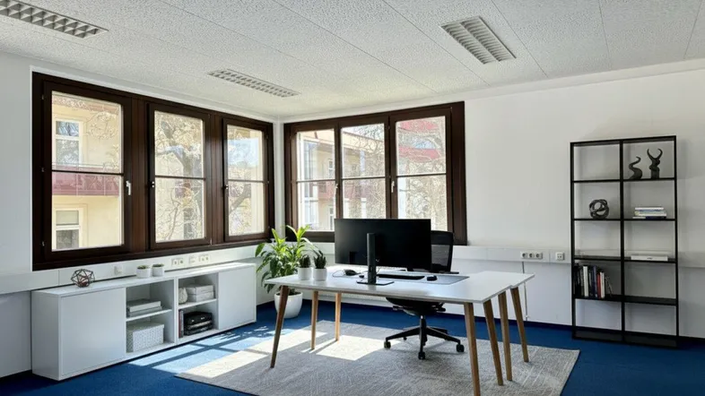 Modernes Büro mit blauer Teppichboden, großen Fenstern und funktionaler Einrichtung für einen Arbeitsplatz.