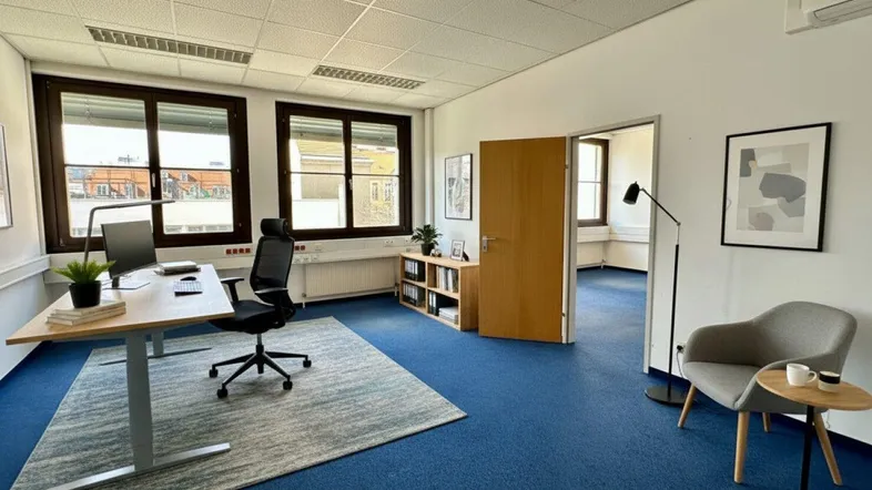 Modernes Büro mit blauem Teppichboden, großen Fenstern und Blick auf die städtische Umgebung.