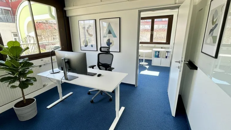Modernes Büro mit blauem Teppichboden, großem Fenster und Blick ins Grüne.