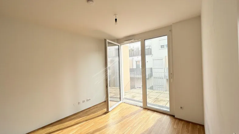 Helles, leeres Zimmer mit Parkettboden und Zugang zu einem Außenbereich.