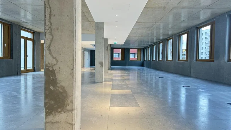 Großzügiger, offener Büroraum mit Sichtbeton, Fliesenboden und vielen Fenstern für natürliches Licht.