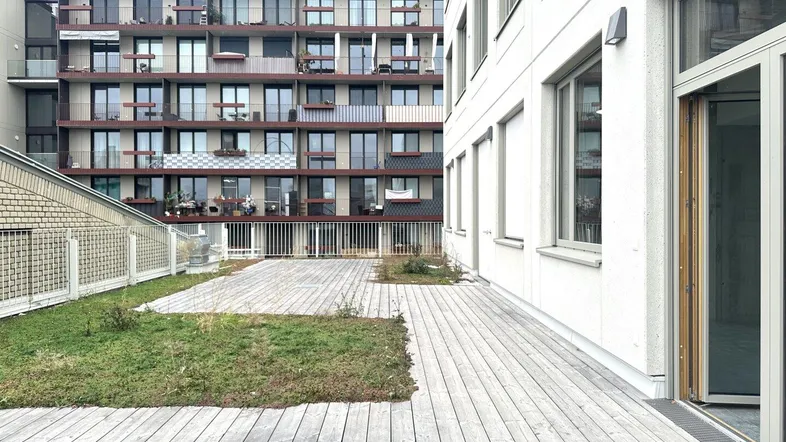 Eine große Holzterrasse mit Grünflächen und Blick auf ein modernes Wohngebäude im Hintergrund.
