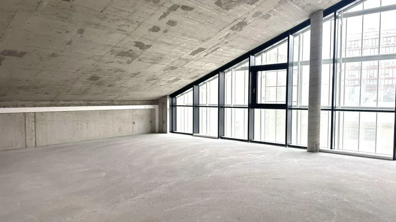 Großer, heller Raum mit Sichtbetondecke und großen, schrägen Fenstern, noch im Rohbauzustand.