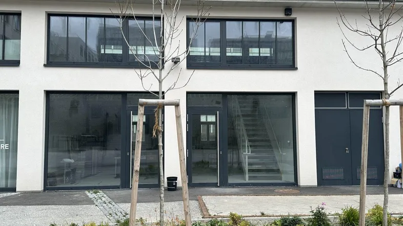 Moderne Gebäudefassade mit großen Schaufenstern und Eingangstüren im Erdgeschoss, umgeben von jungen Bäumen.