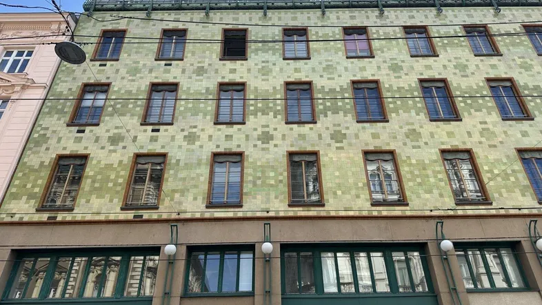 Historische Fassade mit markanten grünen Kacheln und großen Fenstern im Erdgeschoss, die auf eine kommerzielle Nutzung hindeuten.