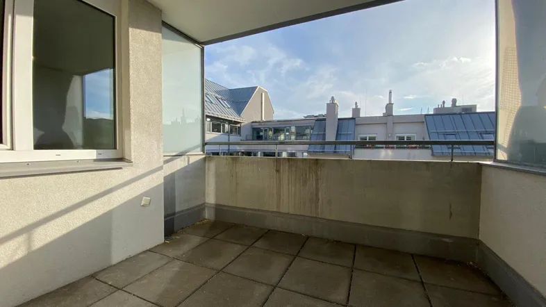 Heller Balkon mit Fliesenboden und Glasbrüstung, bietet einen Blick auf umliegende Gebäude.