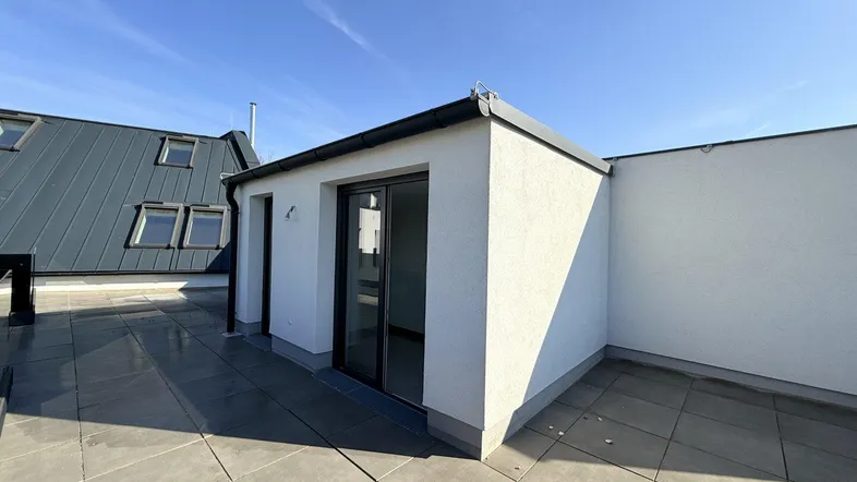 Geräumige Dachterrasse mit modernen Fliesen und Zugang zu einem kleinen Gebäude unter blauem Himmel.