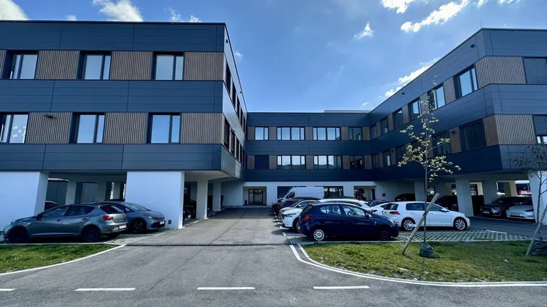 Modernes Gebäude mit dunkler Fassade und Holzdetails, umgeben von Parkplätzen unter blauem Himmel.