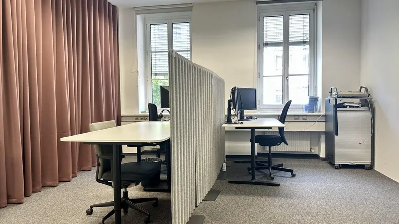 Modernes Büro mit zwei Arbeitsplätzen, Teppichboden und großen Fenstern für viel Tageslicht.