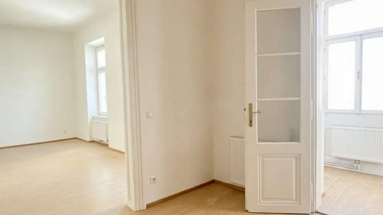 Heller Raum mit Holzboden und weiß gestrichenen Wänden, durch eine Tür mit einem weiteren Raum verbunden.