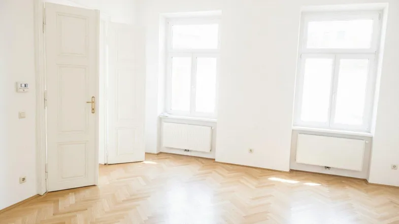 Heller Raum mit Fischgrätparkett und zwei großen Fenstern, die viel Tageslicht hereinlassen.
