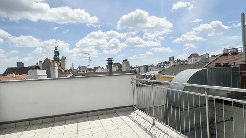 Geräumige Dachterrasse mit Fliesenboden und weitem Blick über die Dächer der Stadt unter blauem Himmel.