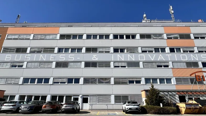 Modernes Geschäftsgebäude mit mehreren Etagen und großen Fenstern, davor ein Parkplatz für Fahrzeuge.