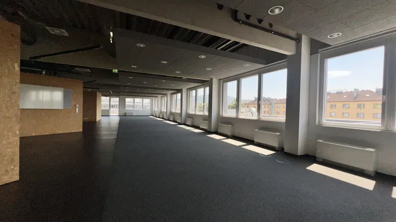 Geräumiger, offener Bürobereich mit großen Fenstern, dunklem Boden und Blick auf die Stadt.