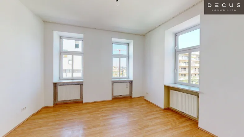 Helles, leeres Zimmer mit Holzfußboden und mehreren Fenstern, die viel Tageslicht hereinlassen.