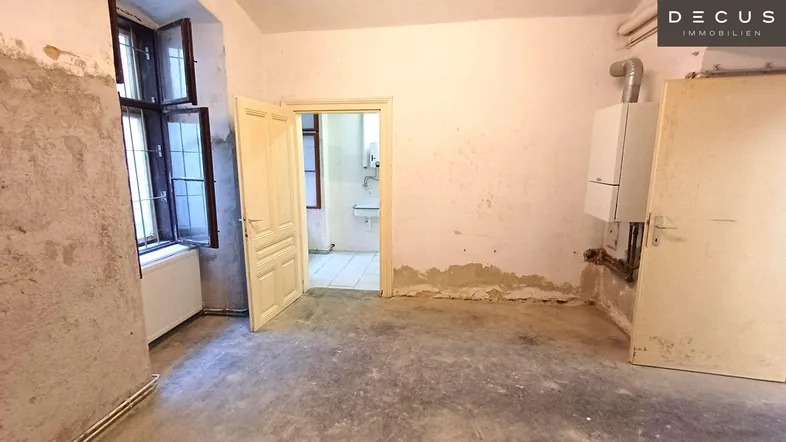 Innenraum in sanierungsbedürftigem Zustand mit freigelegten Wänden, alten Fenstern und einem Heizkessel.