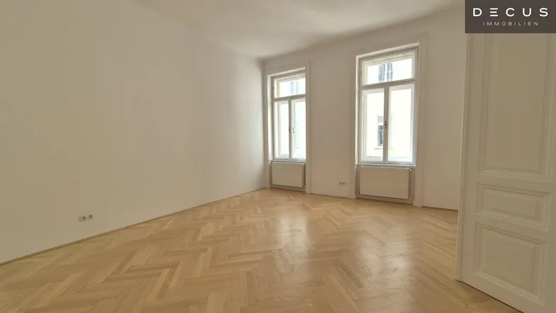 Helles, leeres Zimmer mit Fischgrätparkettboden und hohen Fenstern, bereit zur individuellen Gestaltung.