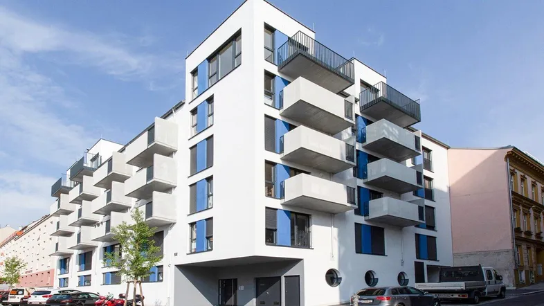 Modernes Mehrfamilienhaus mit weißer Fassade, blauen Akzenten und zahlreichen Balkonen in urbaner Umgebung.