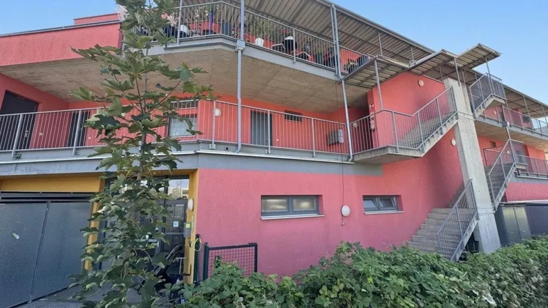 Modernes Mehrfamilienhaus mit leuchtend roter Fassade, Balkonen und Außentreppen, umgeben von Grün.