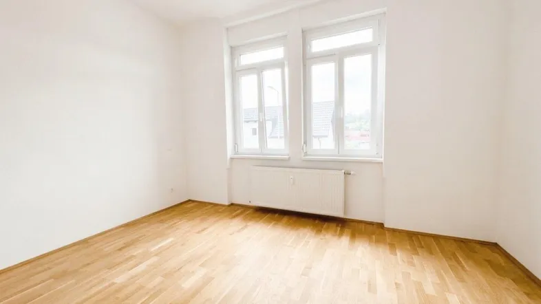 Helles Zimmer mit Parkettboden und großen Fenstern, ideal als Wohn- oder Schlafbereich nutzbar.