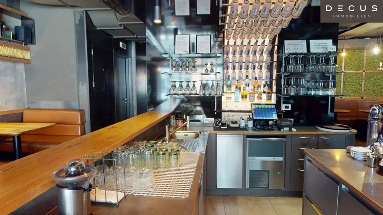 Moderne Bar mit Holztheke, vielen Gläsern und Flaschen, sowie Sitzgelegenheiten im Hintergrund.