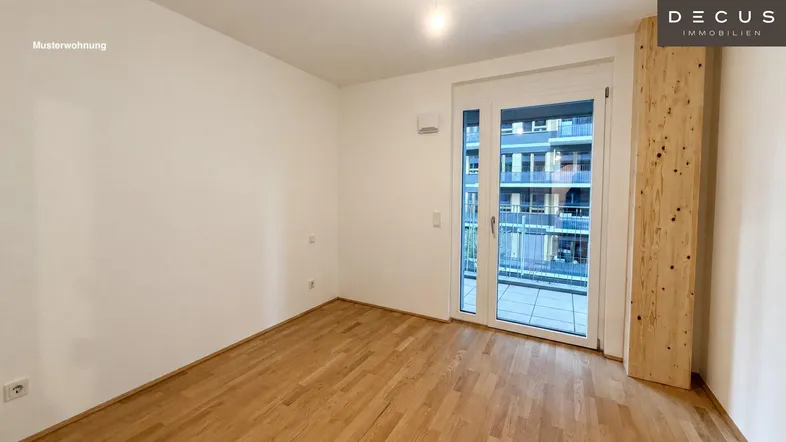 Helles, leeres Zimmer mit Holzboden und Zugang zu einem Balkon in einer Musterwohnung.