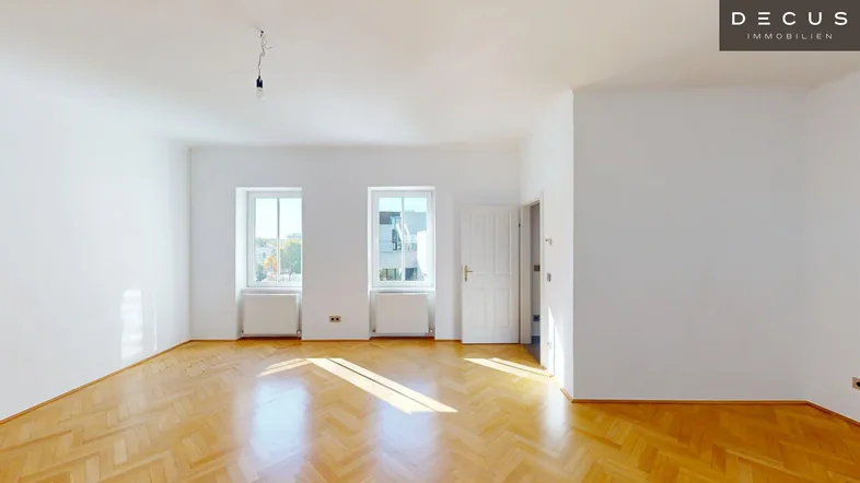 Helles, leeres Zimmer mit Fischgrätparkettboden und zwei Fenstern, das viel Tageslicht hereinlässt.