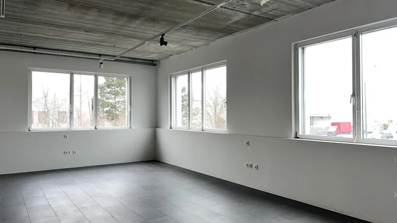 Leerer Büroraum mit großen Fenstern, Fliesenboden und einer modernen Sichtbetondecke.