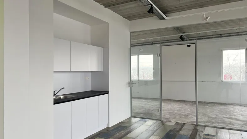 Modernes Büro mit kleiner Küchenzeile, freiliegender Betondecke und Glasabtrennung zu einem weiteren Raum.